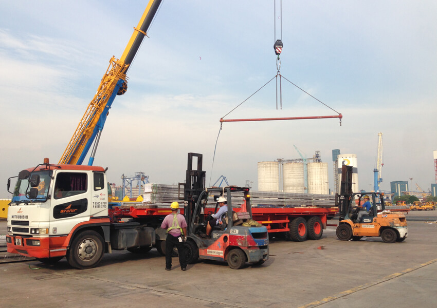 Crane and forklifts loading heavy equipment onto an Avenir Logistics truck at a port.